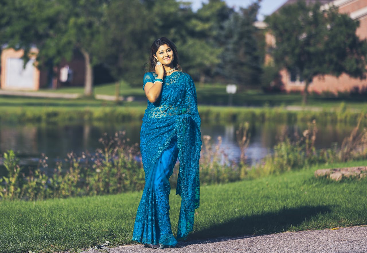 An elegant woman in a blue saree poses by a tranquil park pond on a sunny day.
