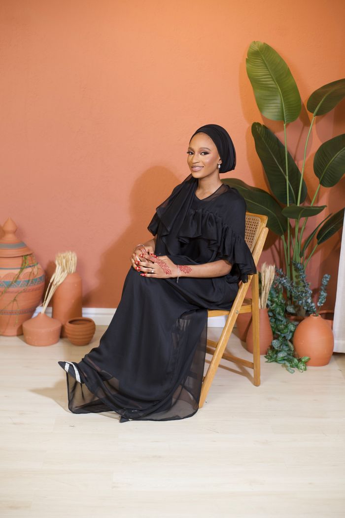 Stylish woman in a black dress and headwrap sitting indoors, showcasing elegance and cultural fashion.